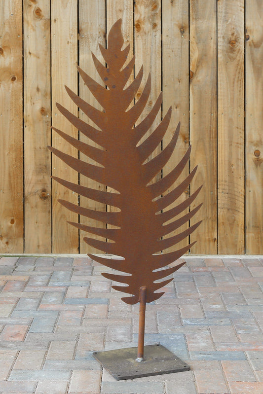 Decorative metal fern leaf sculpture on a stand against a wooden fence.